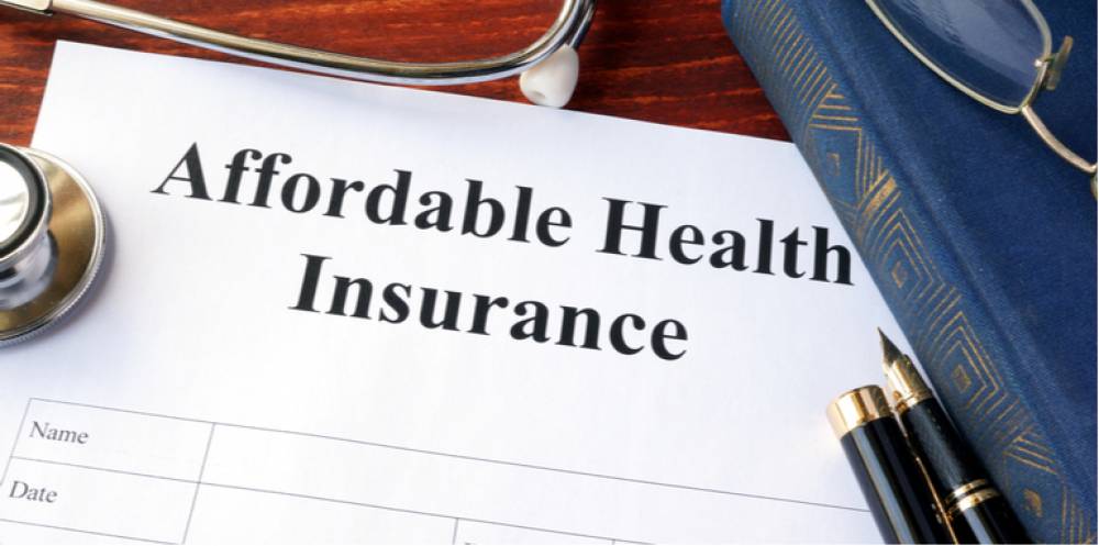  The image shows a stethoscope, a book, a pen, and a pair of glasses on a table. The paper on the table has 'Affordable Health Insurance' written on it.
